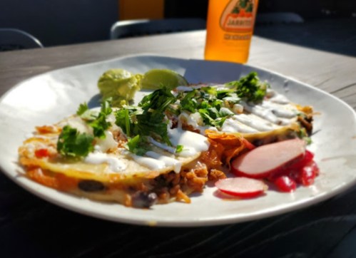 A plate of tacos topped with cilantro, sour cream, and lime, served with radishes and a bottle of Jarritos soda.