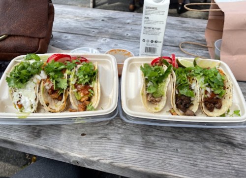 Two containers of tacos with various fillings, topped with cilantro and onions, on a wooden table.