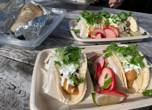 Two tacos topped with cilantro and sauce, served with radishes and lime, alongside a wrapped burrito in a container.