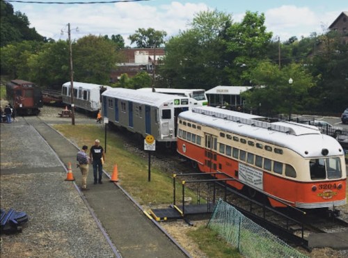 A vintage streetcar and modern train cars are parked on tracks, with people walking nearby in a green, scenic area.