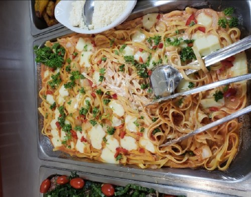 A tray of pasta topped with cheese, diced tomatoes, and parsley, with serving utensils beside it.