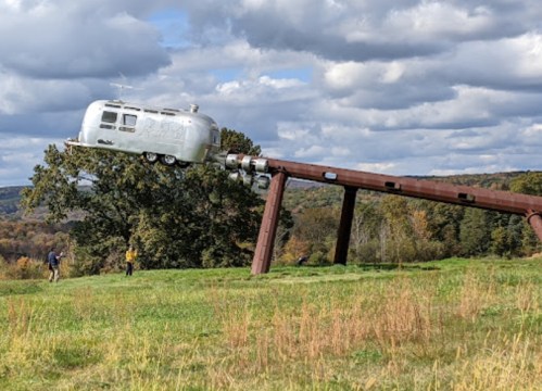 A silver Airstream trailer is suspended at an angle on a large metal structure in a grassy landscape with trees and clouds.