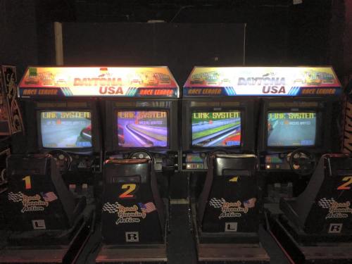 Four arcade racing game machines with screens displaying "Daytona USA" and linked gameplay options.