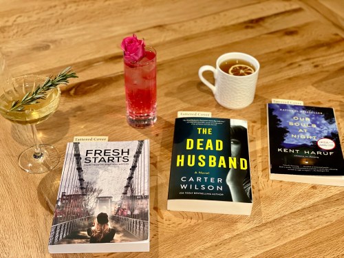 A wooden table with three books, a cocktail, a pink drink, and a cup of tea, creating a cozy reading atmosphere.