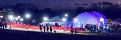 A festive outdoor event with tents, lights, and a dome, surrounded by people and trees at dusk.