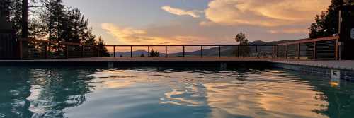 A serene pool at sunset, reflecting colorful clouds and surrounded by trees and mountains in the background.