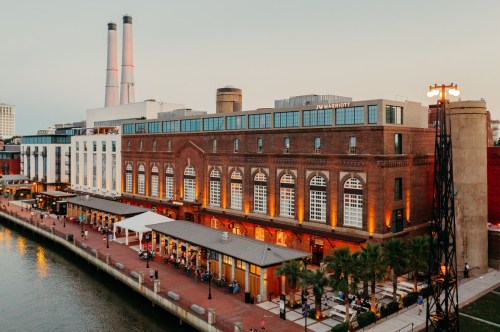 A waterfront view of a brick building with large windows, featuring outdoor seating and two tall smokestacks in the background.