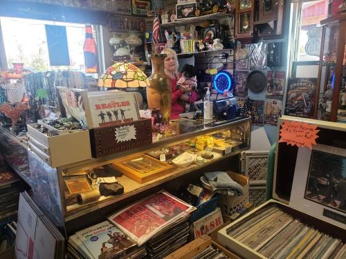 A cluttered antique shop filled with various collectibles, records, and a woman standing behind the counter.