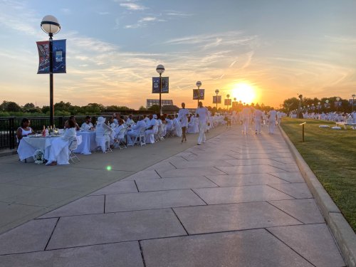 A sunset view of a white-themed outdoor dining event with guests seated at tables along a walkway.