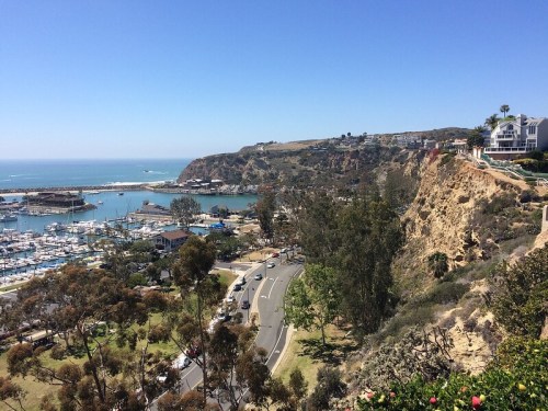A scenic view of a coastal marina with boats, cliffs, and clear blue skies. Lush greenery lines the road.