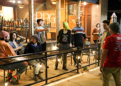 A group of people socializing outside a restaurant at night, with chairs stacked inside and string lights overhead.