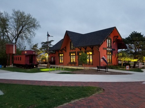 A vibrant orange train station building with a red train car nearby, set in a park with trees and pathways.