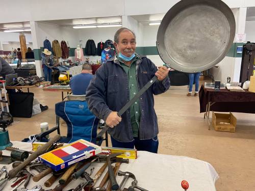 A smiling man holds a large frying pan in a market filled with various tools and items for sale.