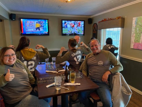 A group of people in matching shirts pose at a table, smiling and showing off their shirt numbers in a cozy bar setting.