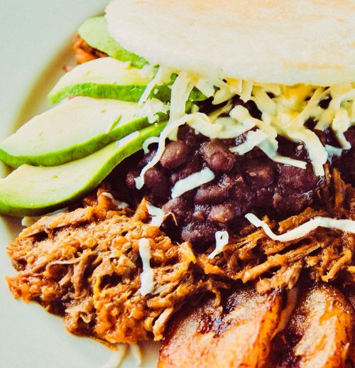 A plate of shredded meat, black beans, avocado slices, cheese, and fried plantains, served with a corn arepa.