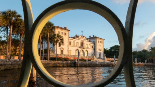 A historic mansion framed by a circular railing, reflecting in calm water with palm trees in the background.