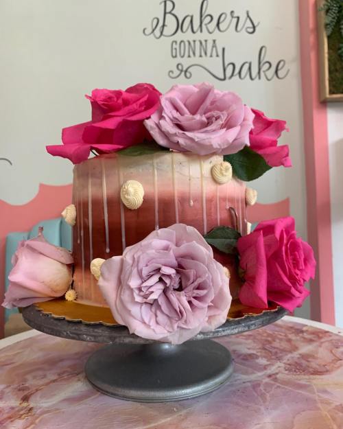 A beautifully decorated cake with pink roses on top, featuring a gradient design and drizzled icing, displayed on a stand.