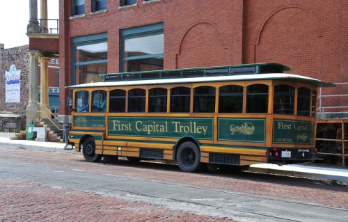 A vintage-style trolley bus parked beside a brick building, featuring "First Capital Trolley" signage.