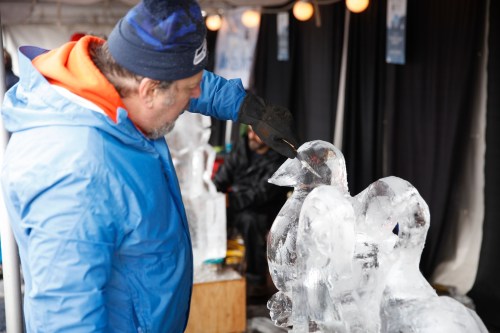 A man carves an ice sculpture of a bird at an outdoor event, wearing a blue jacket and gloves.