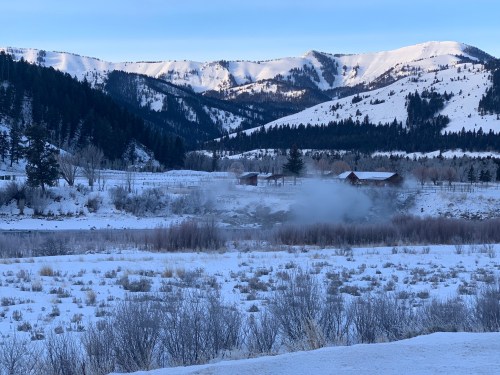 A snowy landscape with mountains in the background, a river, and a few buildings surrounded by trees.