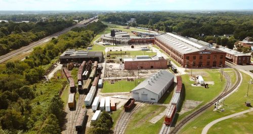 Aerial view of a railway yard with trains, buildings, and green spaces under a clear blue sky.