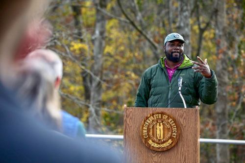 A man in a green jacket and cap speaks at a podium with the Kentucky state seal, surrounded by trees.