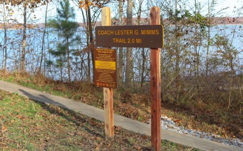 Sign for Coach Lester G. Mimms Trail, indicating a 2.0-mile path near a lake, surrounded by trees and grass.