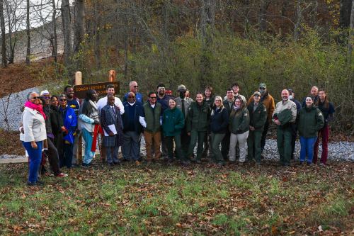 A diverse group of people stands together in front of a sign in a wooded area, smiling for the camera.