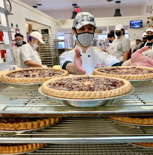 A baker in a kitchen presents freshly baked pecan pies on a cooling rack, with other bakers in the background.