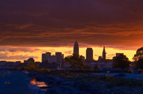 Silhouette of a city skyline at sunset, with dark clouds and vibrant orange and purple hues over the water.