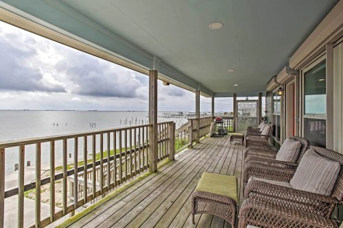 A spacious porch with wicker chairs overlooks a calm waterway under a cloudy sky.