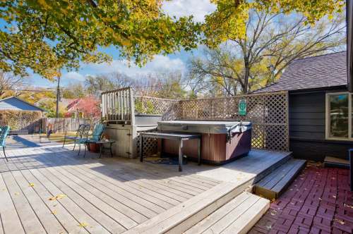 A wooden deck with a hot tub, surrounded by trees and outdoor seating in a sunny backyard.