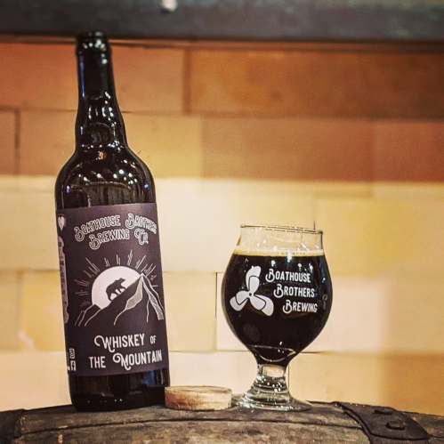 A bottle of "Whiskey of the Mountain" and a glass on a wooden barrel, featuring Boathouse Brothers Brewing branding.