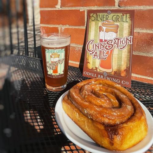 A cinnamon roll on a plate next to a glass of beer, with a sign for "Cinnsation Ale" in the background.