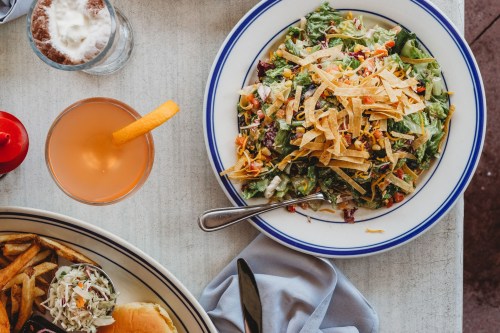 A colorful salad topped with crispy tortilla strips, served with a drink and side of fries on a table.
