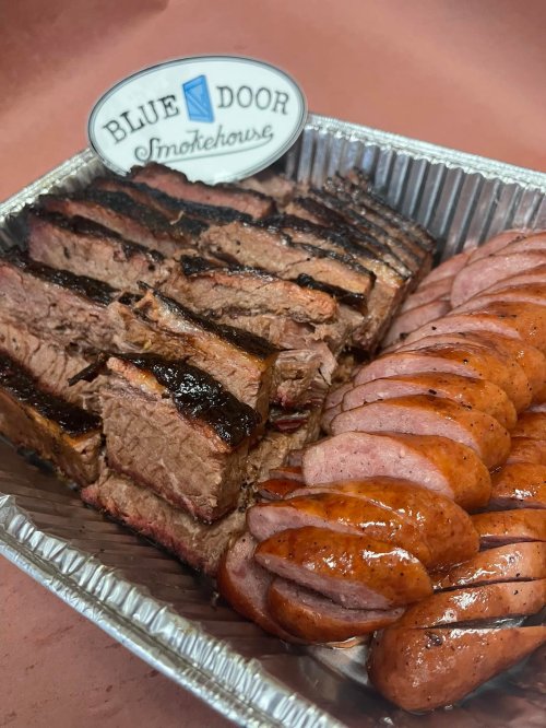 A tray of sliced smoked brisket and sausage, with a "Blue Door Smokehouse" sign on top.