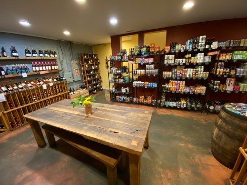 A cozy wine and beer shop featuring wooden shelves, a large table, and a variety of bottles and cans on display.
