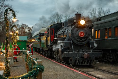 A vintage steam train with smoke billowing, surrounded by festive decorations and people in holiday attire.