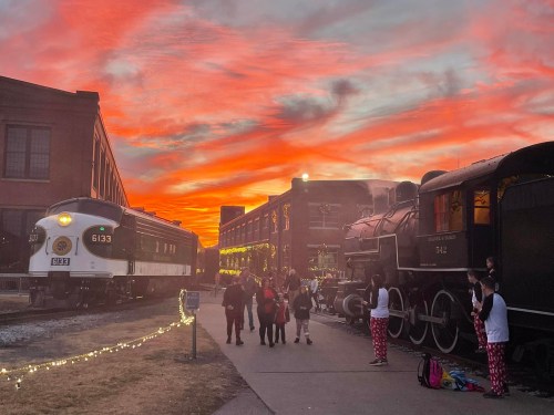 A vibrant sunset casts orange and pink hues over a train station with vintage trains and people gathered nearby.