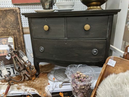 A vintage black dresser with two drawers, gold knobs, and decorative items around it, including dried flowers.