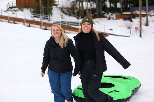 Two women walk joyfully on snow, wearing winter clothing, with a green snow tube beside them.