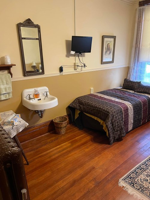 Cozy room with a bed, wall-mounted TV, sink, mirror, and a window, featuring warm wooden floors and decorative elements.