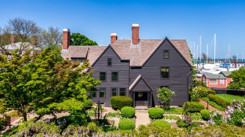 A historic black house surrounded by greenery, with a clear blue sky and boats in the background.