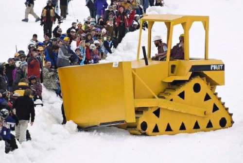 A large yellow bulldozer clears snow while a crowd watches, surrounded by a snowy landscape.