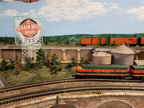 Model train scene featuring a Grain Belt Beer sign, freight cars, and silos amidst greenery and tracks.