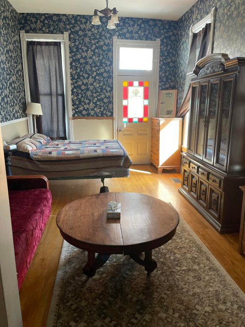 Cozy bedroom with floral wallpaper, a bed, a vintage cabinet, a round table, and a colorful stained glass door.