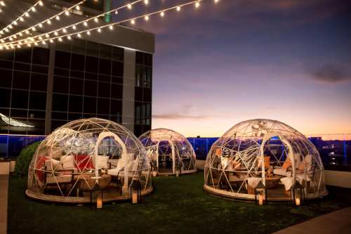 Three transparent domes on a rooftop terrace, illuminated by string lights, with a sunset backdrop.