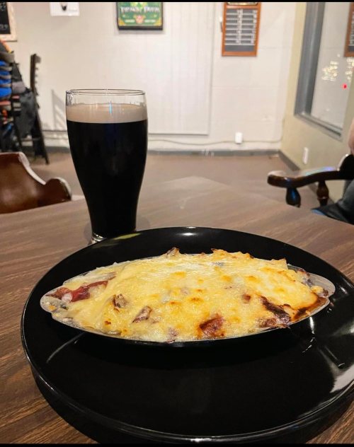 A baked dish topped with melted cheese on a black plate, alongside a glass of dark beer on a wooden table.