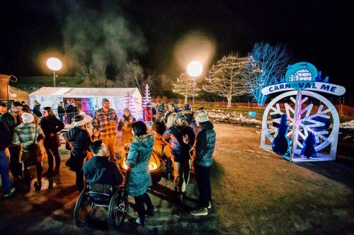 A lively outdoor winter festival scene with people gathered around a fire, festive lights, and a "CARNIVAL ME" sign.