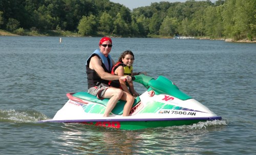 A man and a girl ride a jet ski on a lake, surrounded by trees and calm water on a sunny day.